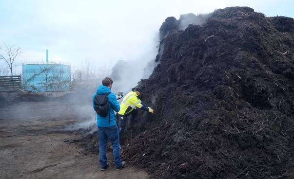 Zwei Männer stehen neben einem riesigen Kompostberg. Der eine nimmt eine Probe vom Kompost, der andere filmt die Situation.